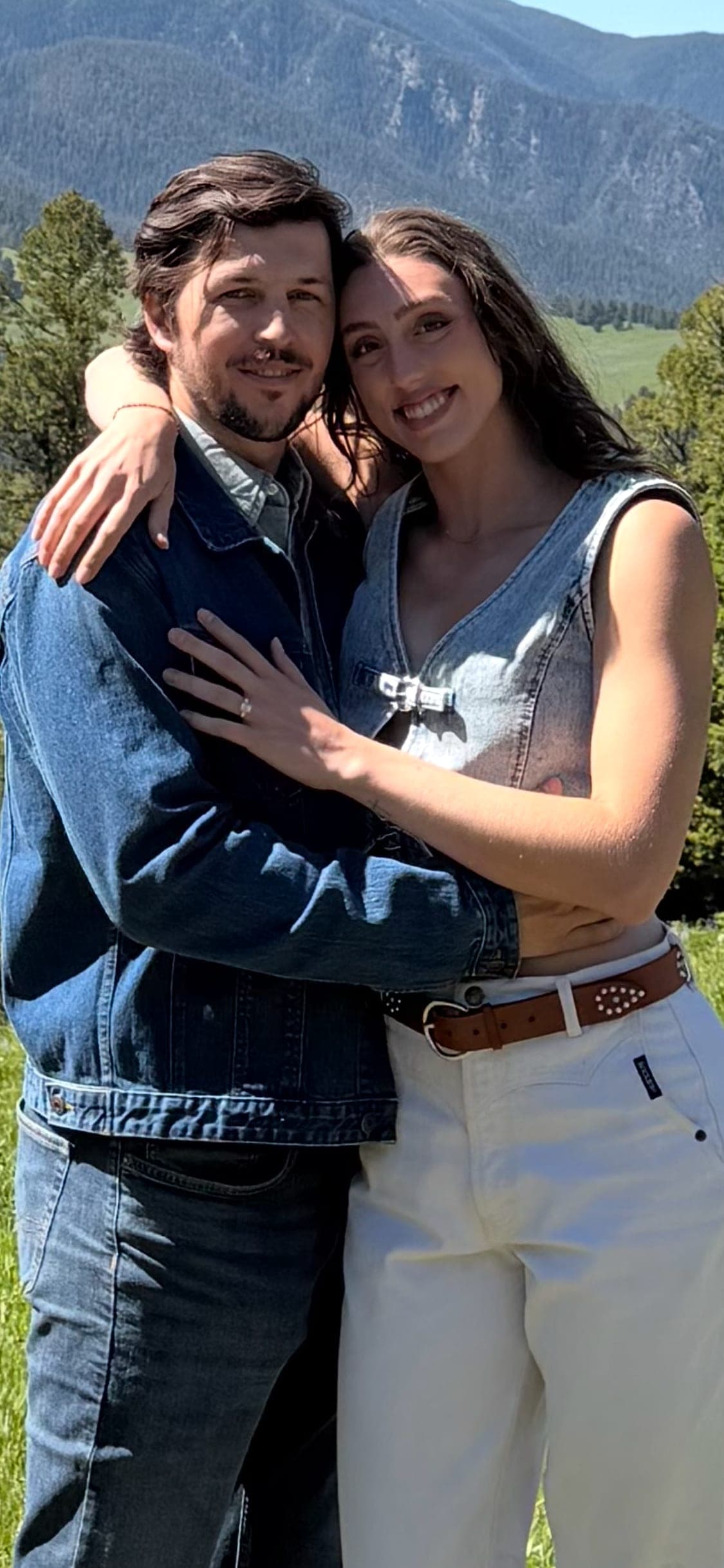 Newly-engaged couple embracing in a mountain meadow — emerald-cut diamond ring on her finger