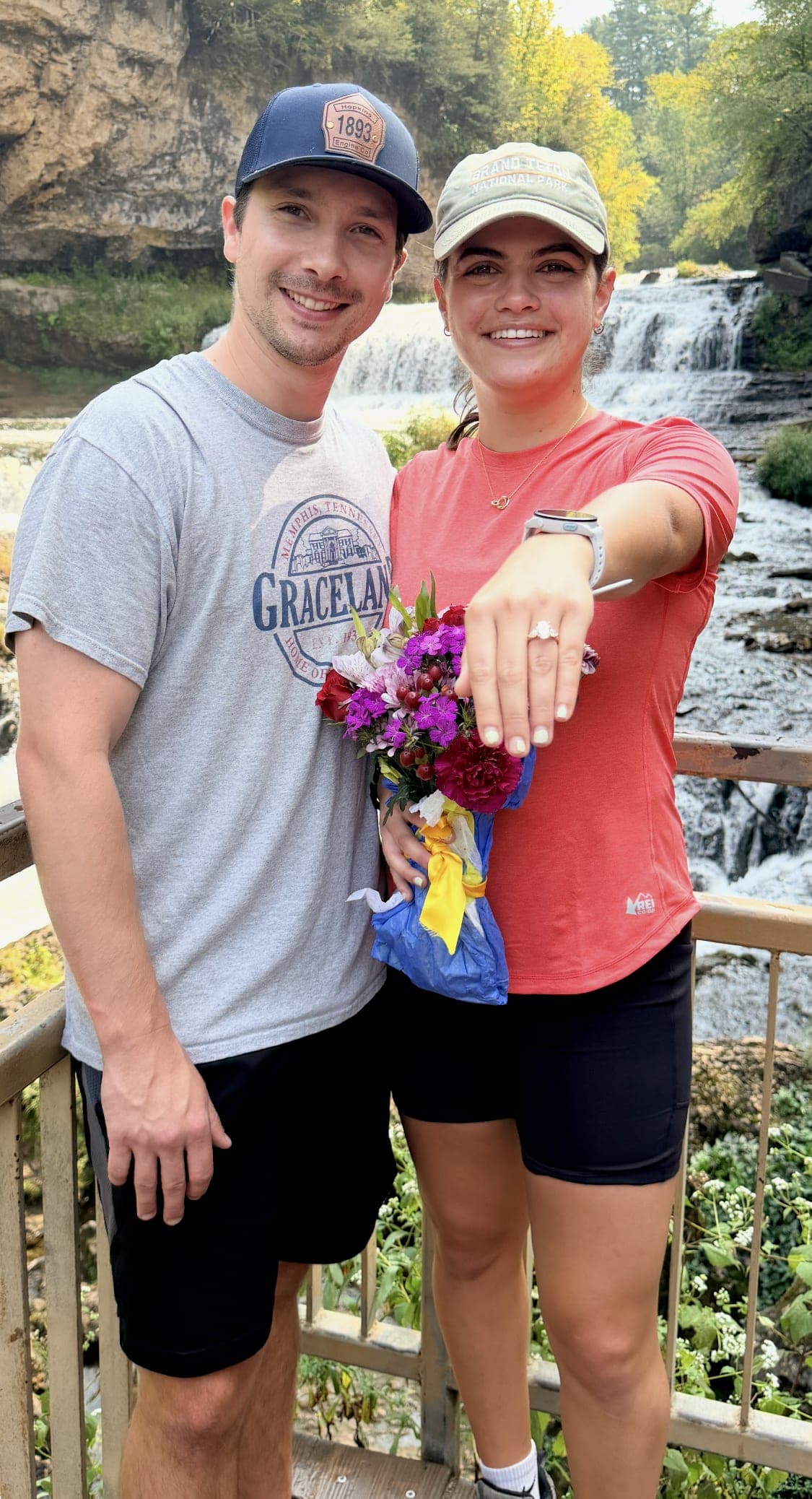 Newly-engaged couple in front of a waterfall — she holds a bouquet and shows off her oval diamond ring